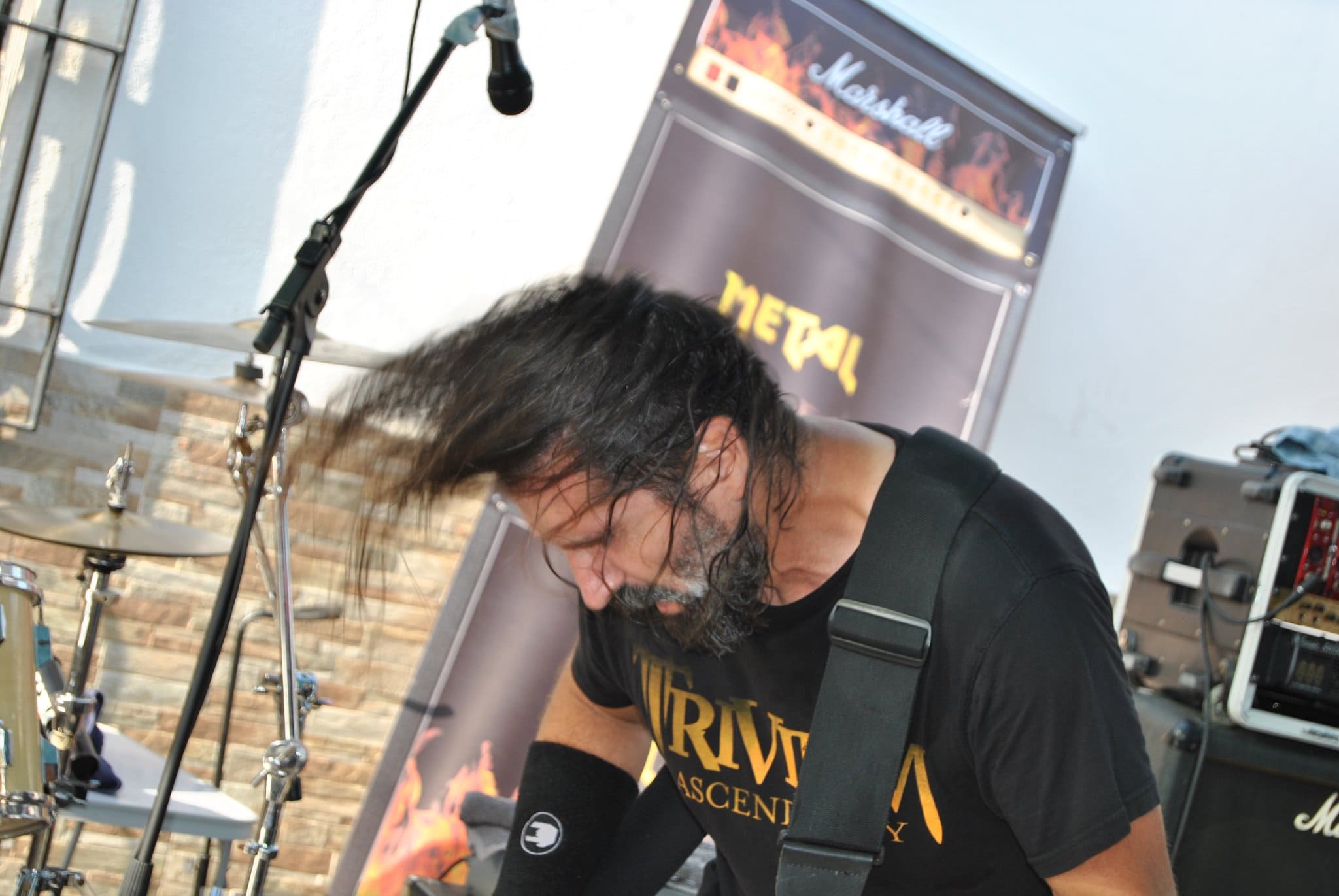 
    La imagen captura el momento enérgico de un guitarrista durante una actuación en vivo. Él está inclinado hacia adelante con su cabeza baja, su cabello largo y oscuro fluye hacia abajo cubriéndole la cara, capturando una sensación de movimiento y pasión por la música. Está vestido con una camiseta negra que lleva la palabra TRIVIUM, que es probablemente el nombre de la banda a la que representa, y se puede ver parte de una camiseta con la palabra METAL detrás de él, probablemente un telón de fondo del escenario. Un amplificador Marshall, conocido en la escena del rock y metal, se vislumbra en el fondo, completando esta escena típica de un concierto de rock.